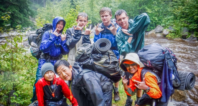 A group of young people wearing backpacks and rain gear pose for a photo beside a river in a densely wooded area. 